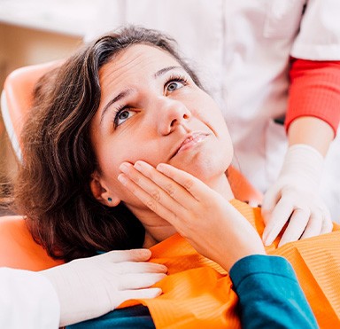 Woman in dental chair touching cheek in pain with 2 dentists touching her shoulders